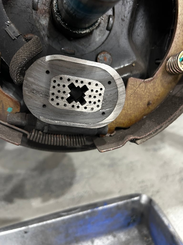Close-up of a worn metal brake component with X-shaped perforations, mounted on rusted automotive parts