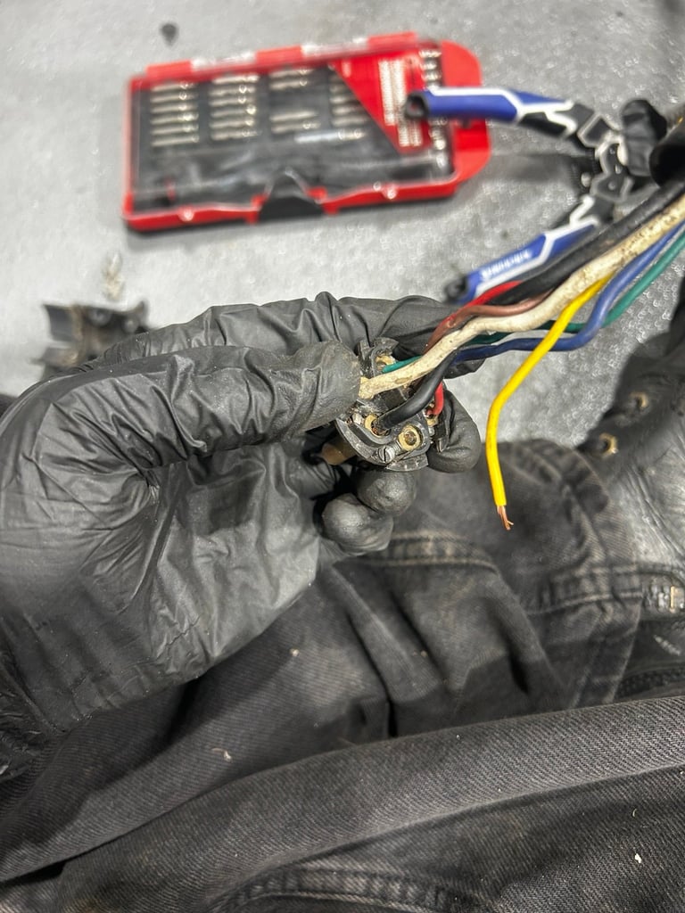 Gloved hand holding car electrical connector with colored wires and red tool kit in background