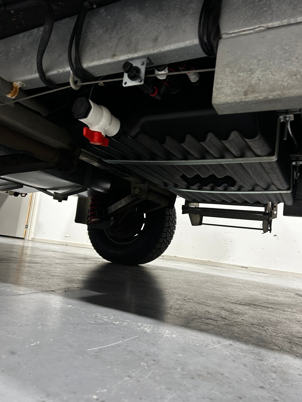 Underside of a truck showing the rear axle, tire, brake light, and undercarriage components on concrete floor