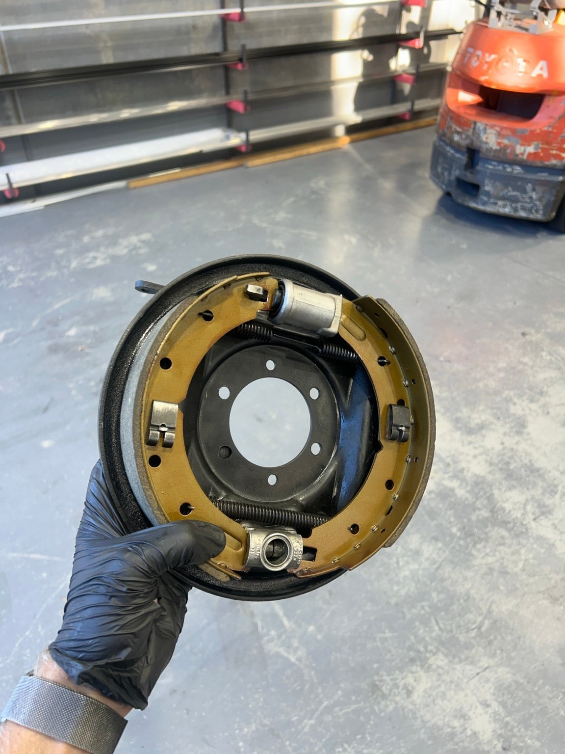 Mechanic in black glove holding a removed brake drum assembly with gold backing plate in garage workshop
