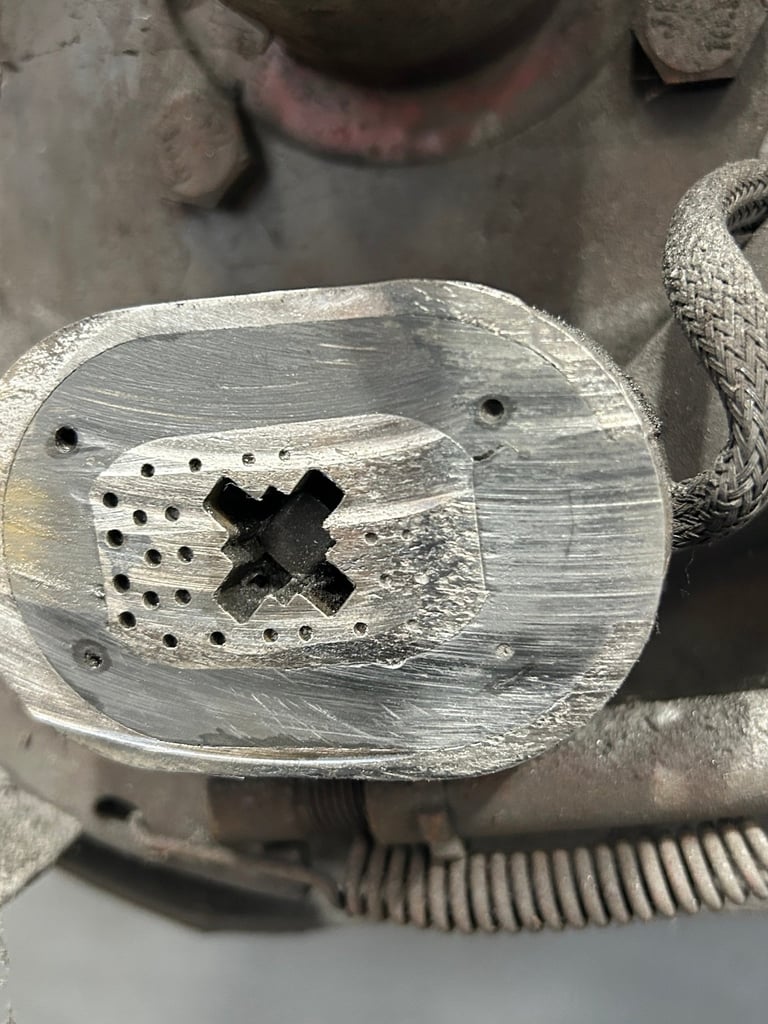 Close-up of a metallic die-cast part with X-shaped hole and circular perforations, mounted in an industrial assembly jig