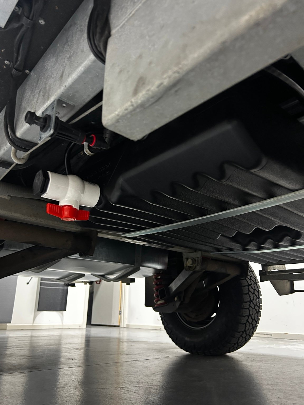 Underside of truck showing black bumper, red taillights, tire, and hydraulic equipment in a garage