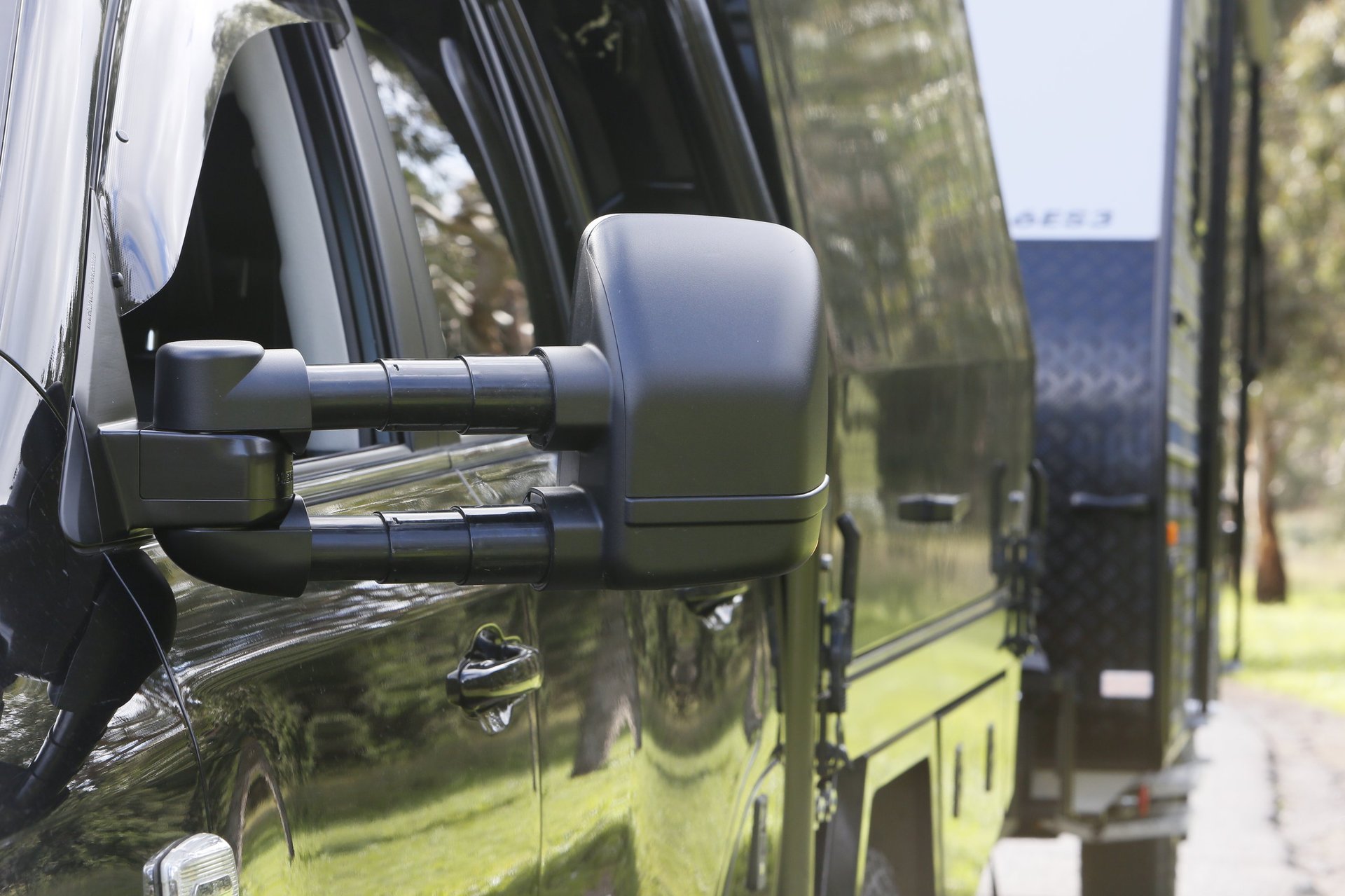Close-up of a white side mirror on a black vehicle with green trees and parked buses visible in the background